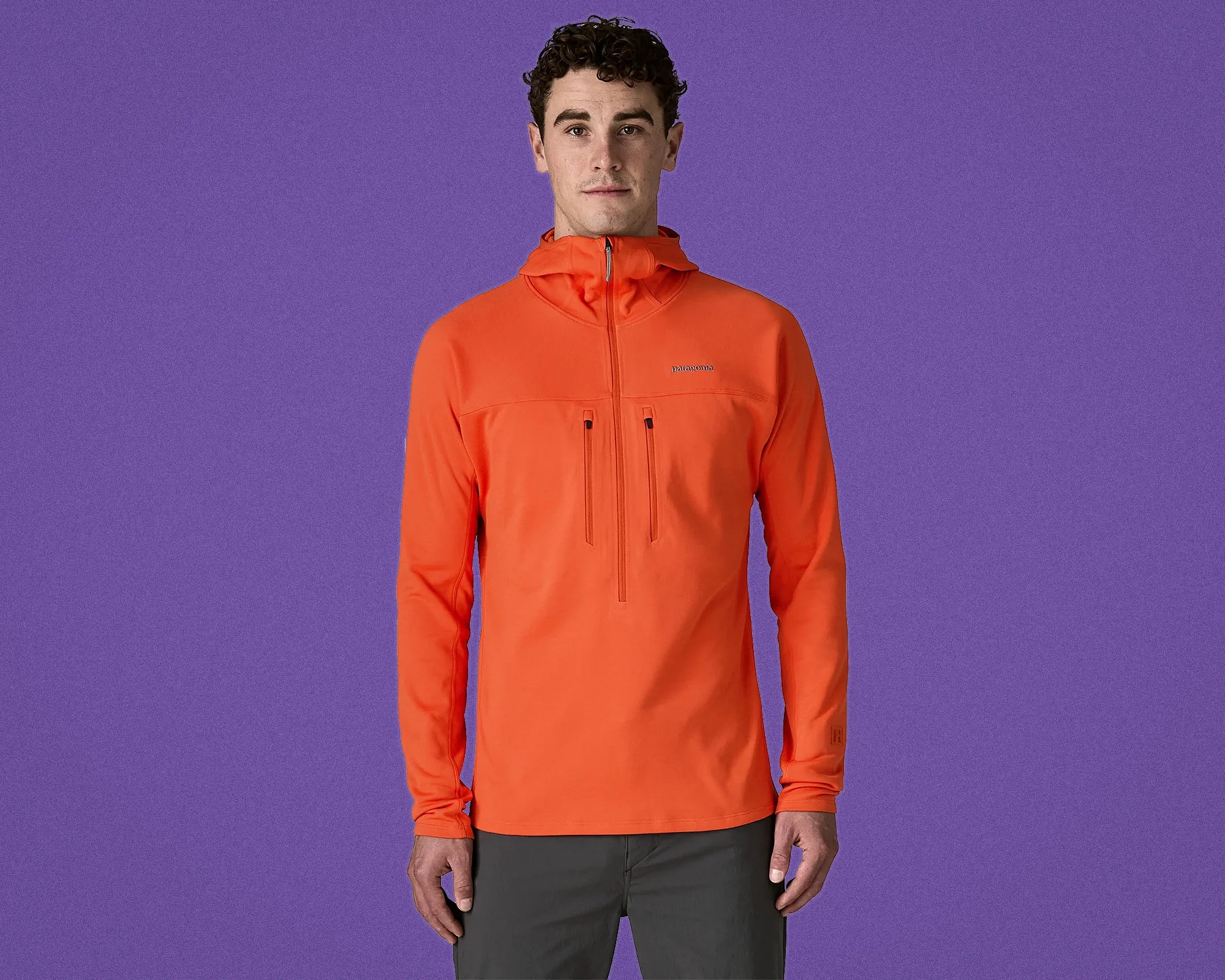 Bright orange long-sleeve hooded pullover with chest zip pockets worn by a person against a purple background.