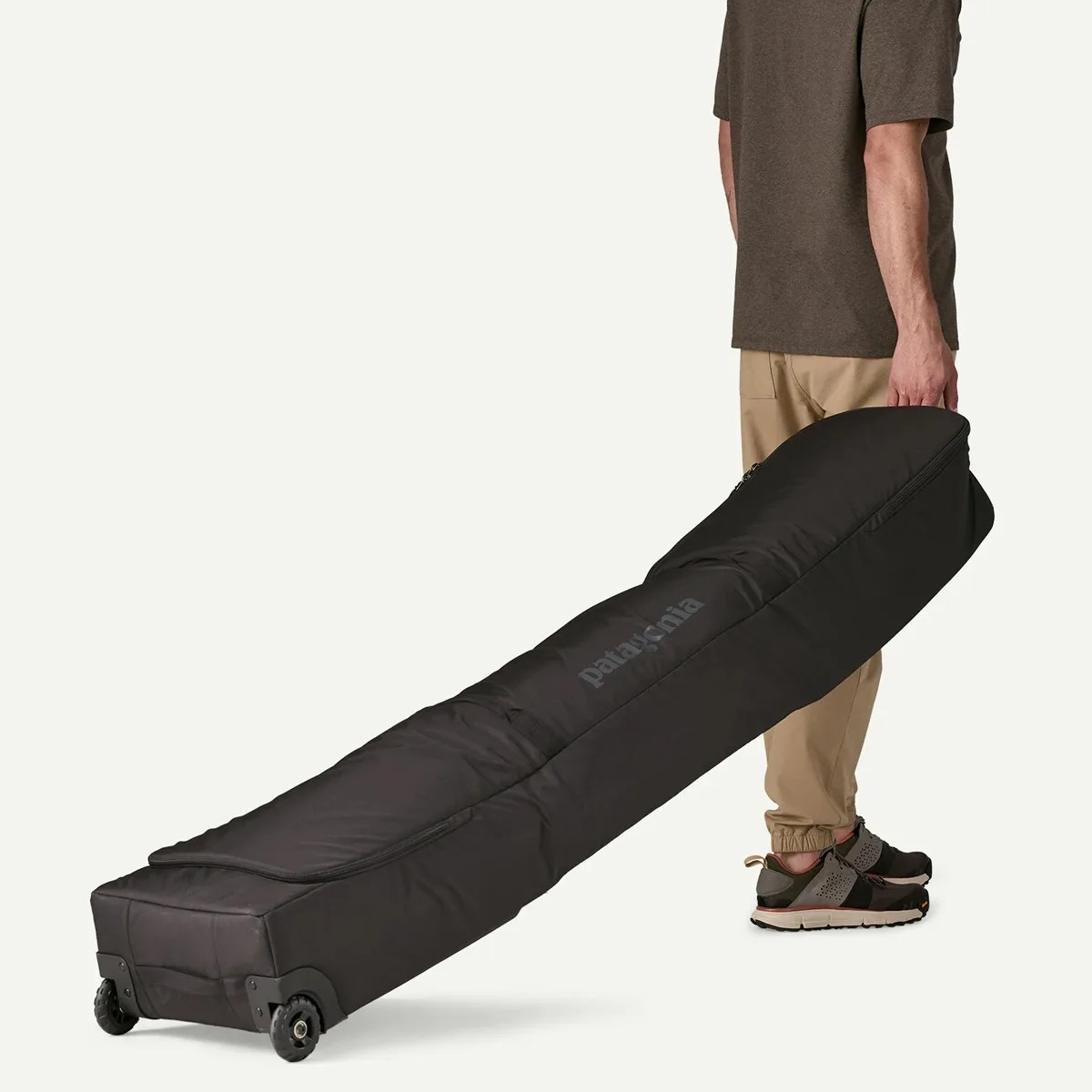 Black wheeled Patagonia gear bag held by a person wearing beige pants and dark sneakers.
