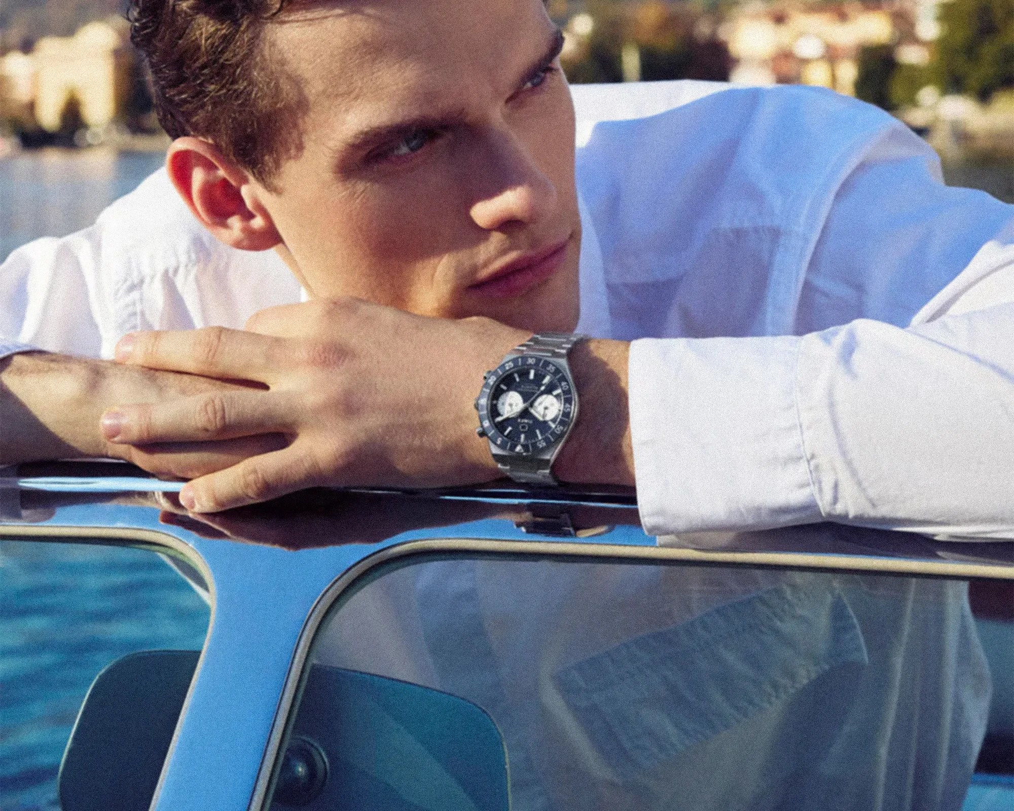 Man in a white shirt leaning on a blue car roof, wearing a silver wristwatch with a black dial.