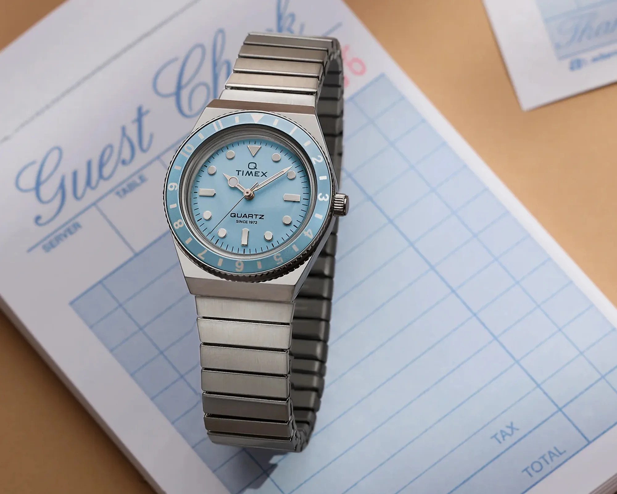 Silver Timex Q Quartz wristwatch with a light blue dial and matching bezel on a guest check pad.