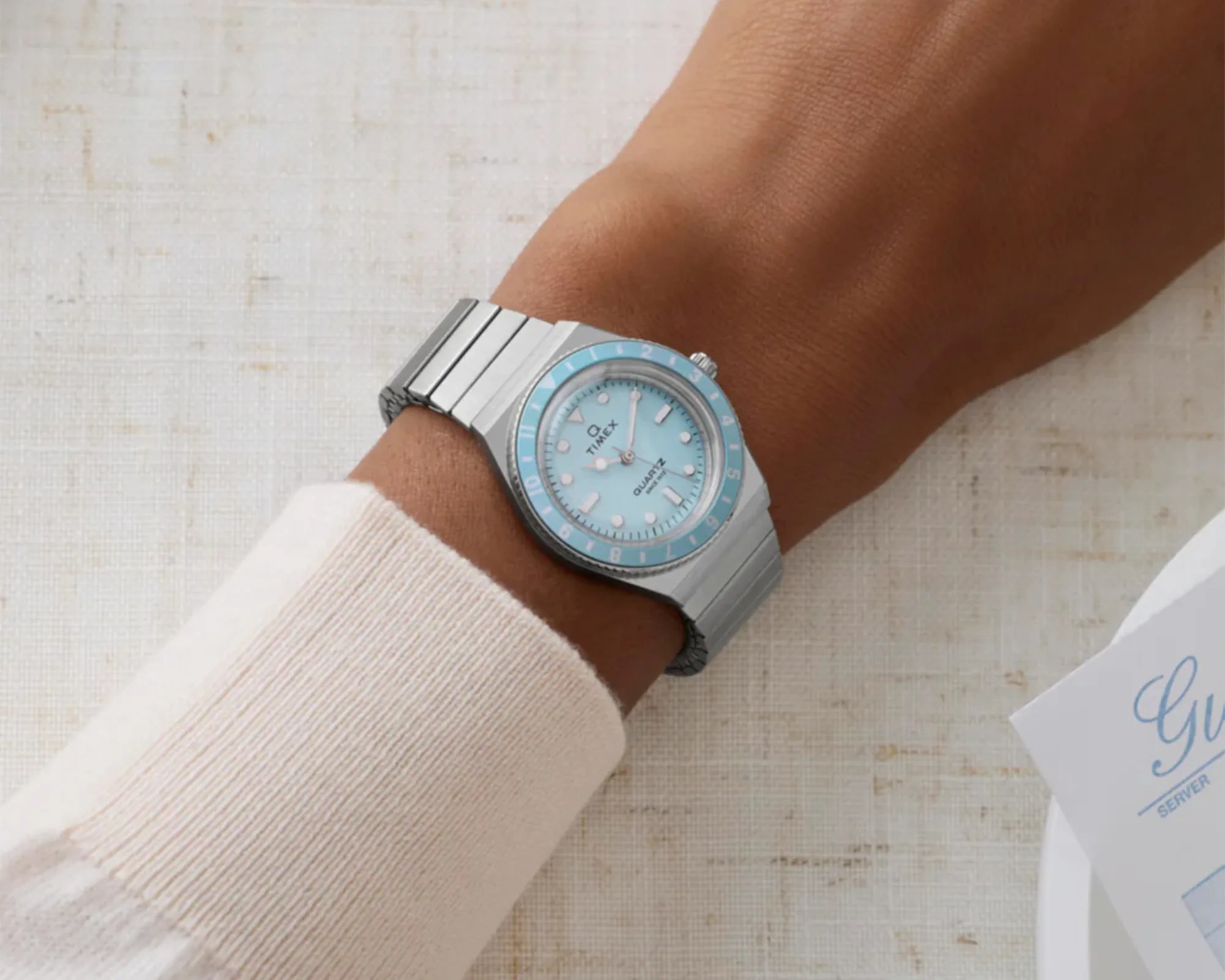 Silver Timex wristwatch with a light blue dial and bezel worn on a wrist with a cream-colored sleeve.