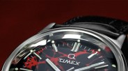 Close-up of a Timex watch face with a black and red abstract pattern and silver hour markers, attached to a black leather strap.