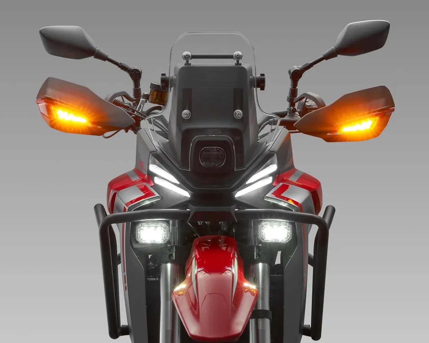 Front view of a red and black motorcycle with illuminated white headlights and orange turn signals on handguards.
