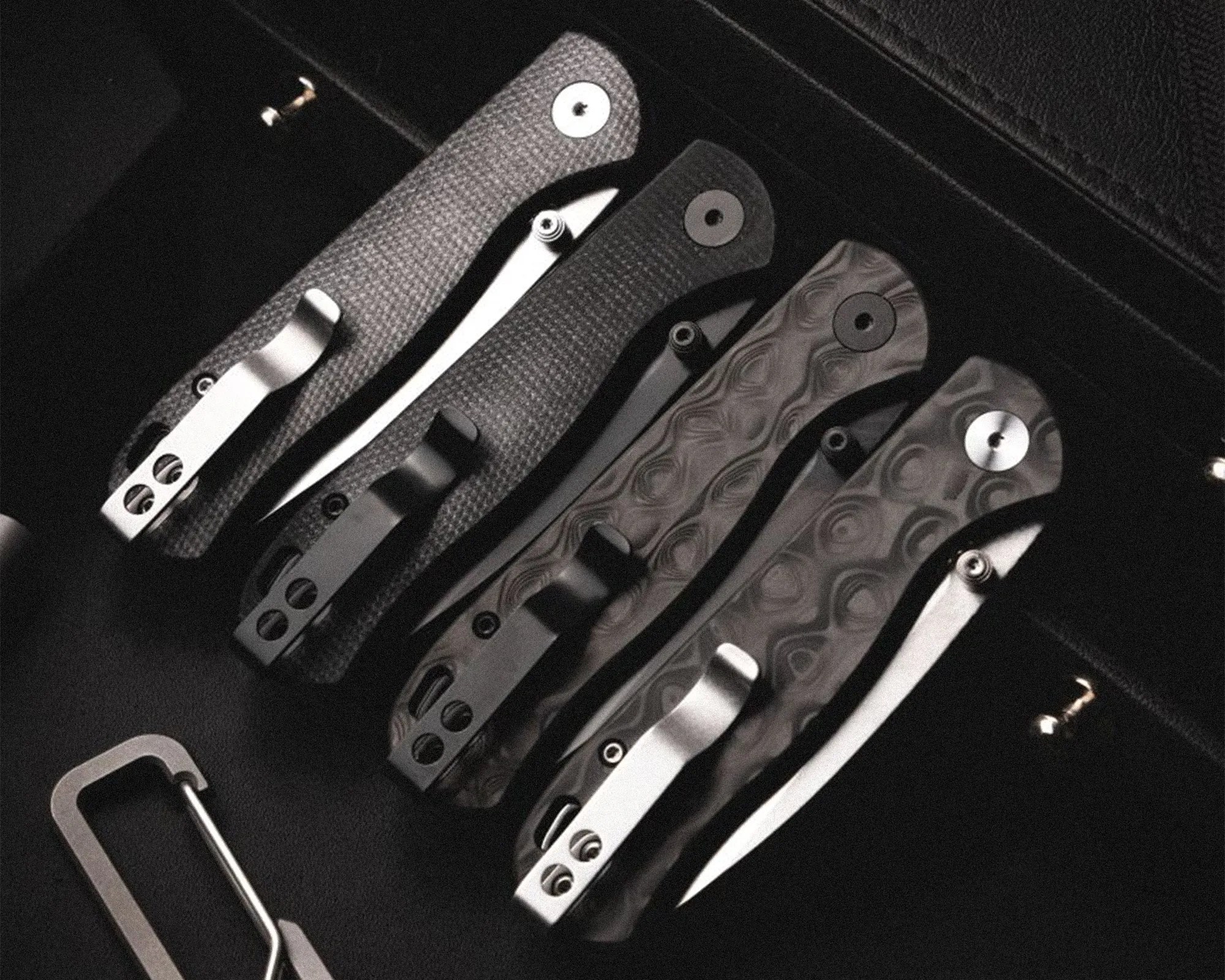 Four folding knives with textured handles in black and dark gray, arranged side by side in a black case.