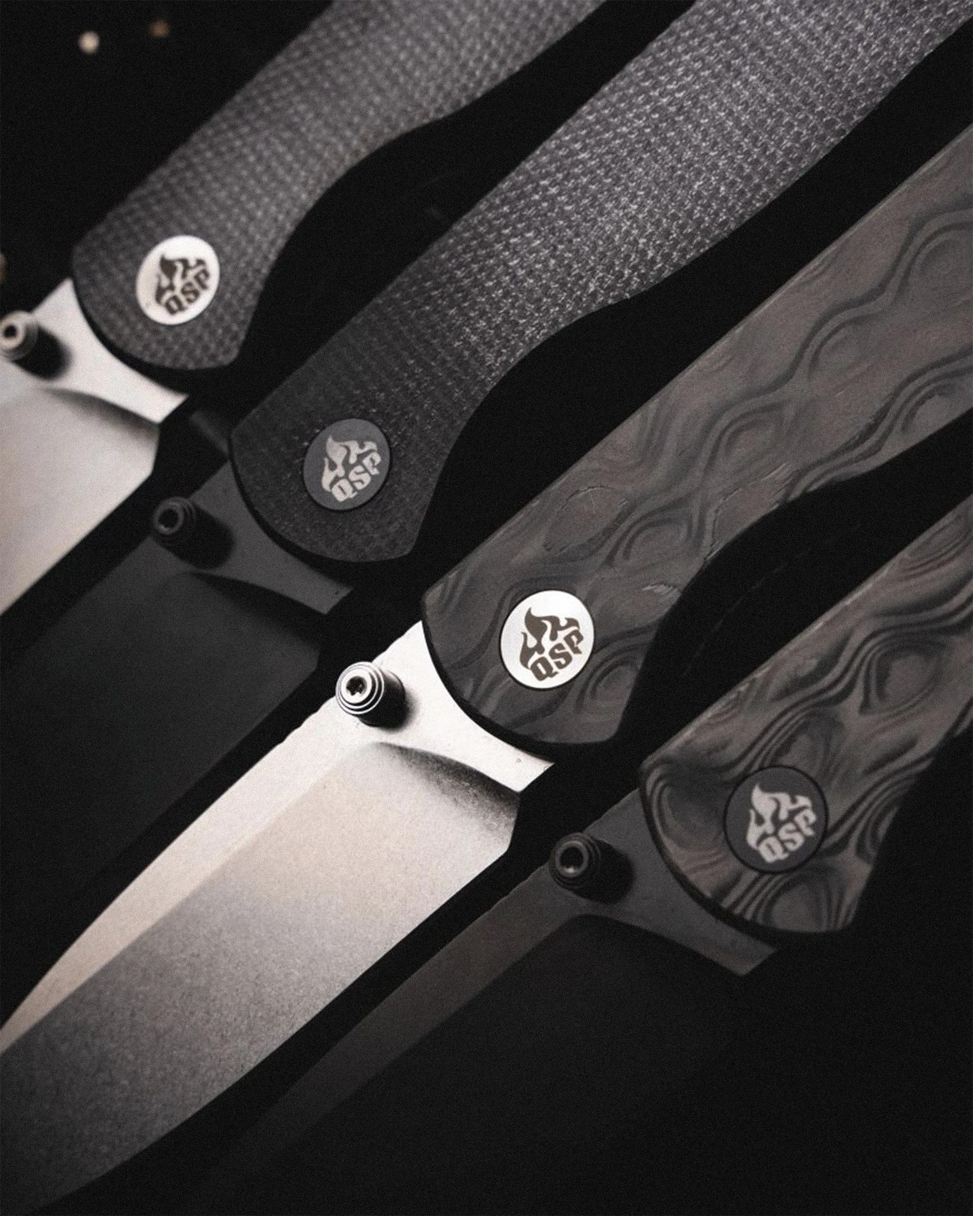 Close-up of four folding knives with textured black handles and QSP logo near the blade pivot.