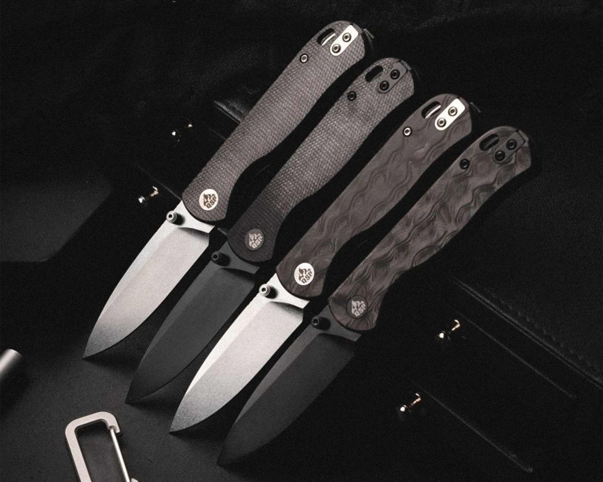 Four folding knives with textured black handles and partially open blades arranged side by side on a dark surface.