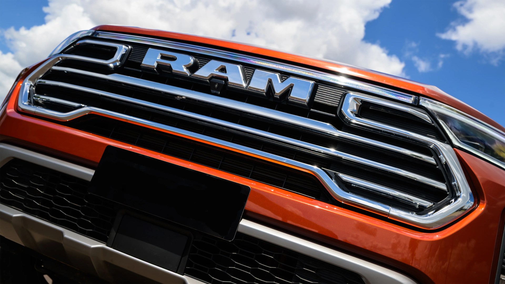 Close-up of an orange RAM truck front grille with chrome accents under a partly cloudy sky.