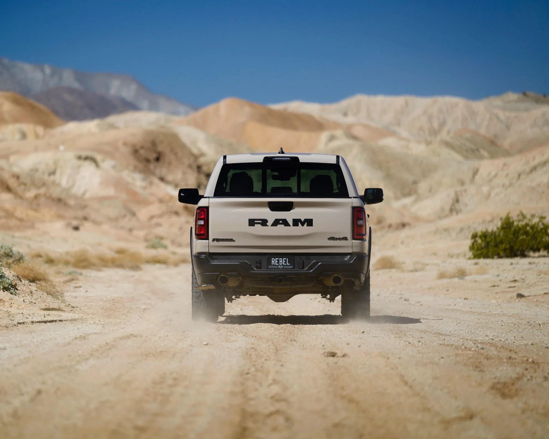 Rear view of a beige RAM Rebel 4x4 pickup truck driving on a dusty desert road with rocky hills in the background.