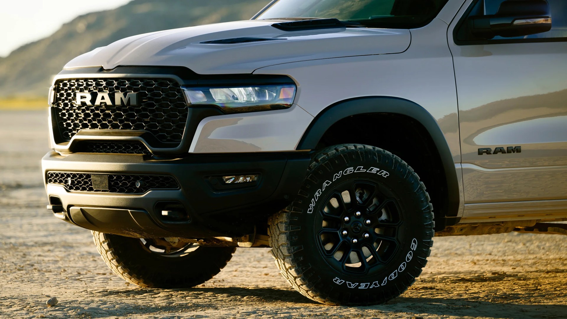 Front side view of a silver RAM truck with black grille and off-road Goodyear Wrangler tires on dry ground.