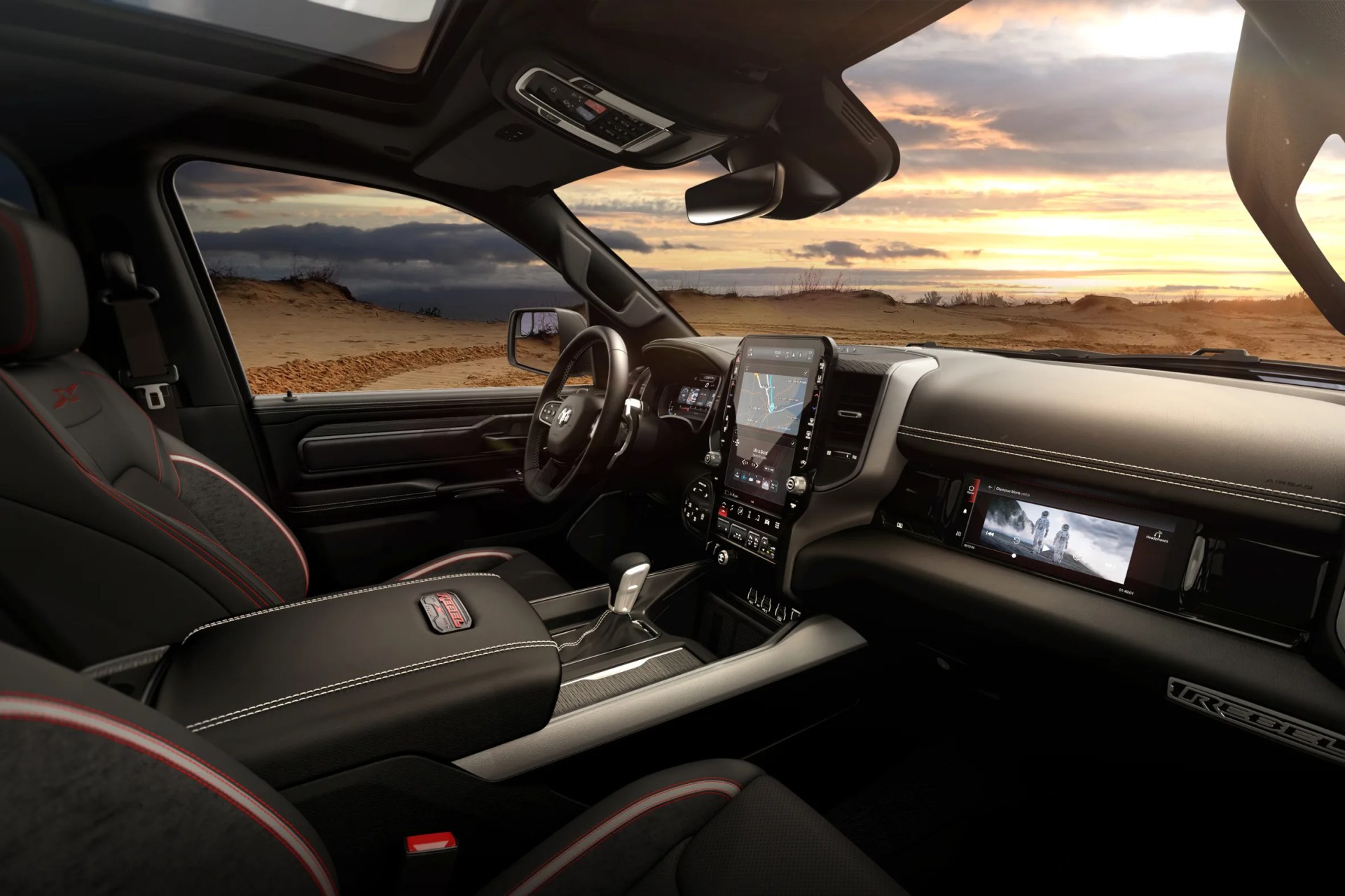Black and red-trimmed truck interior with dual touchscreen displays and leather-trimmed center console at sunset.