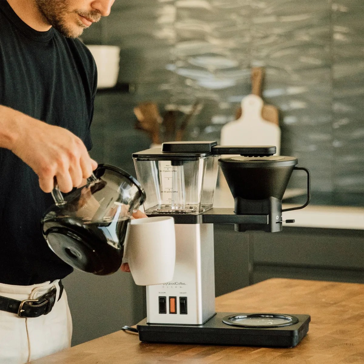 Person pouring coffee from a glass carafe into a white mug next to a black and silver drip coffee maker on a wooden table.