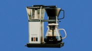 Silver and black drip coffee maker with glass carafe filled with coffee against a blue background.