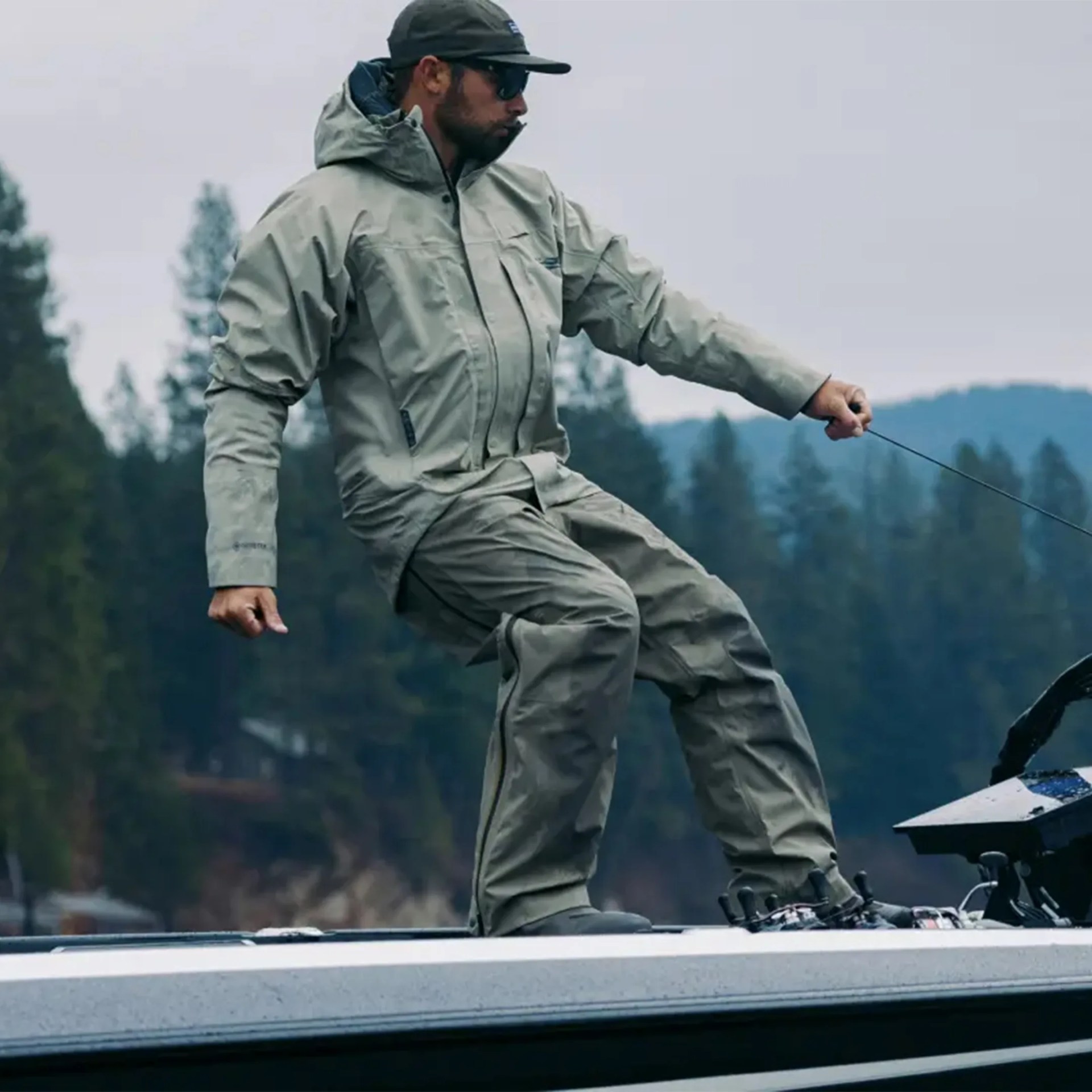 Man wearing a gray waterproof jacket and pants standing on a boat holding a fishing line.