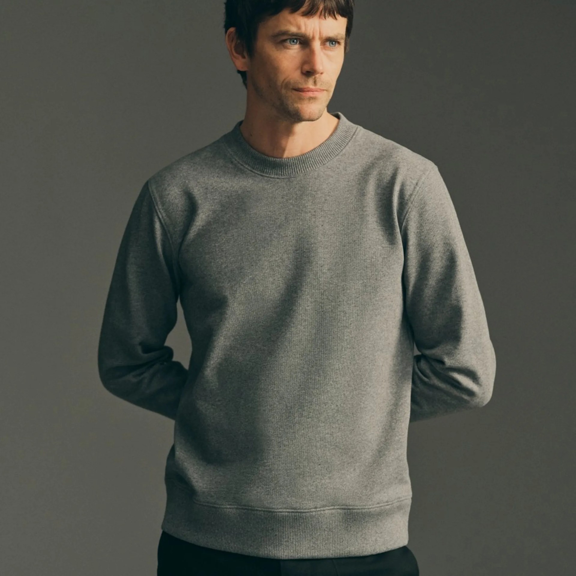 Man wearing a plain grey crewneck sweatshirt with ribbed cuffs and hem against a dark background.