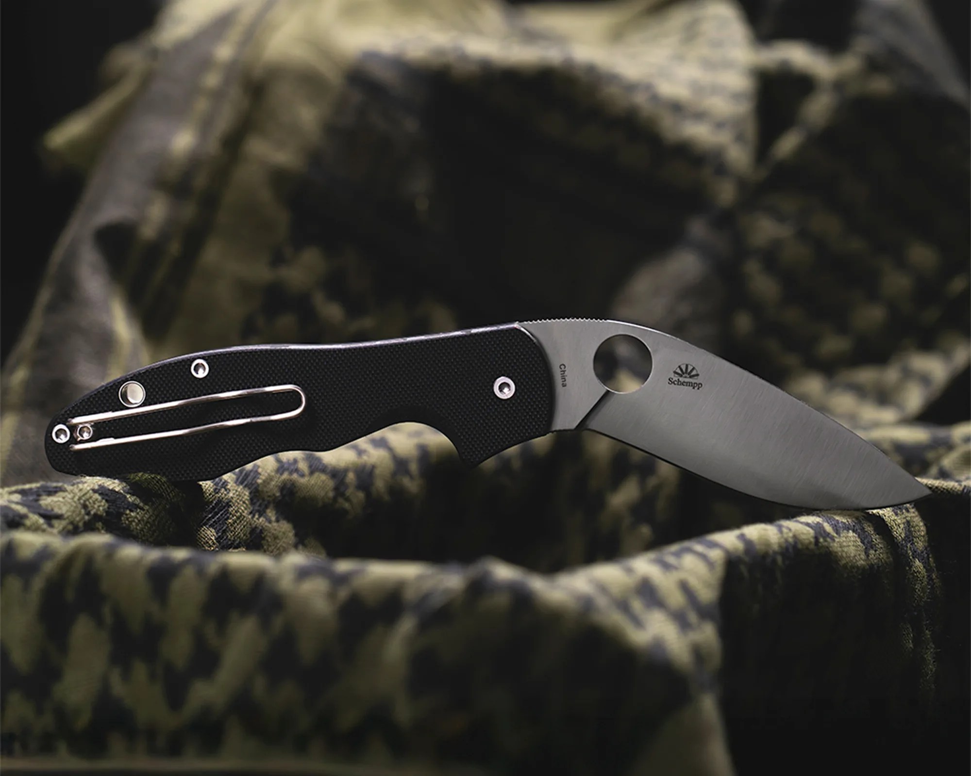 Folding knife with black textured handle and stainless steel blade resting on green and black patterned fabric.