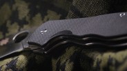 Close-up of a folding knife with textured black handle resting on green and black camouflage fabric.