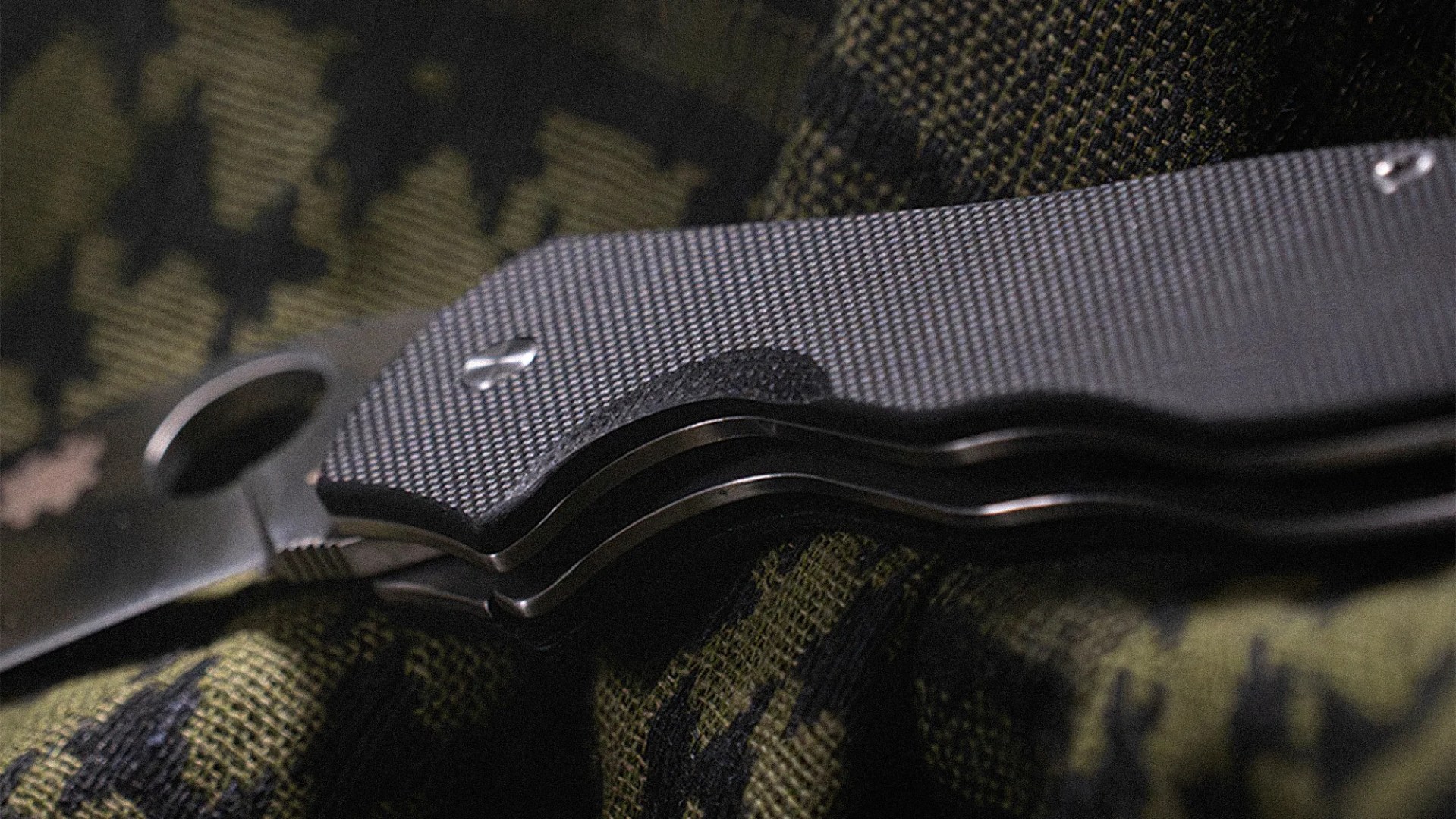 Close-up of a folding knife with textured black handle resting on green and black camouflage fabric.