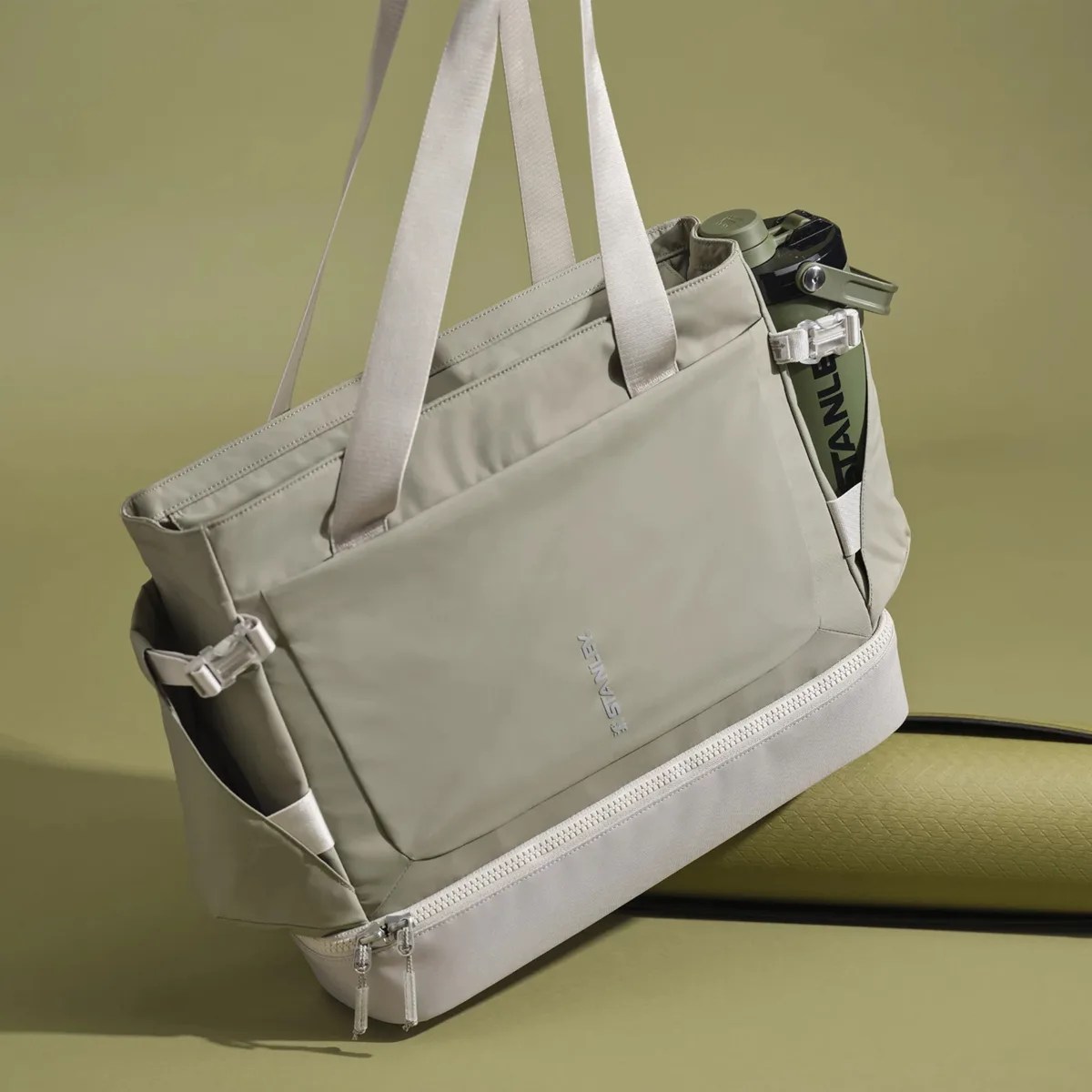 Light gray tote bag with dual handles, side pockets holding a green Stanley bottle, and a zippered bottom compartment.