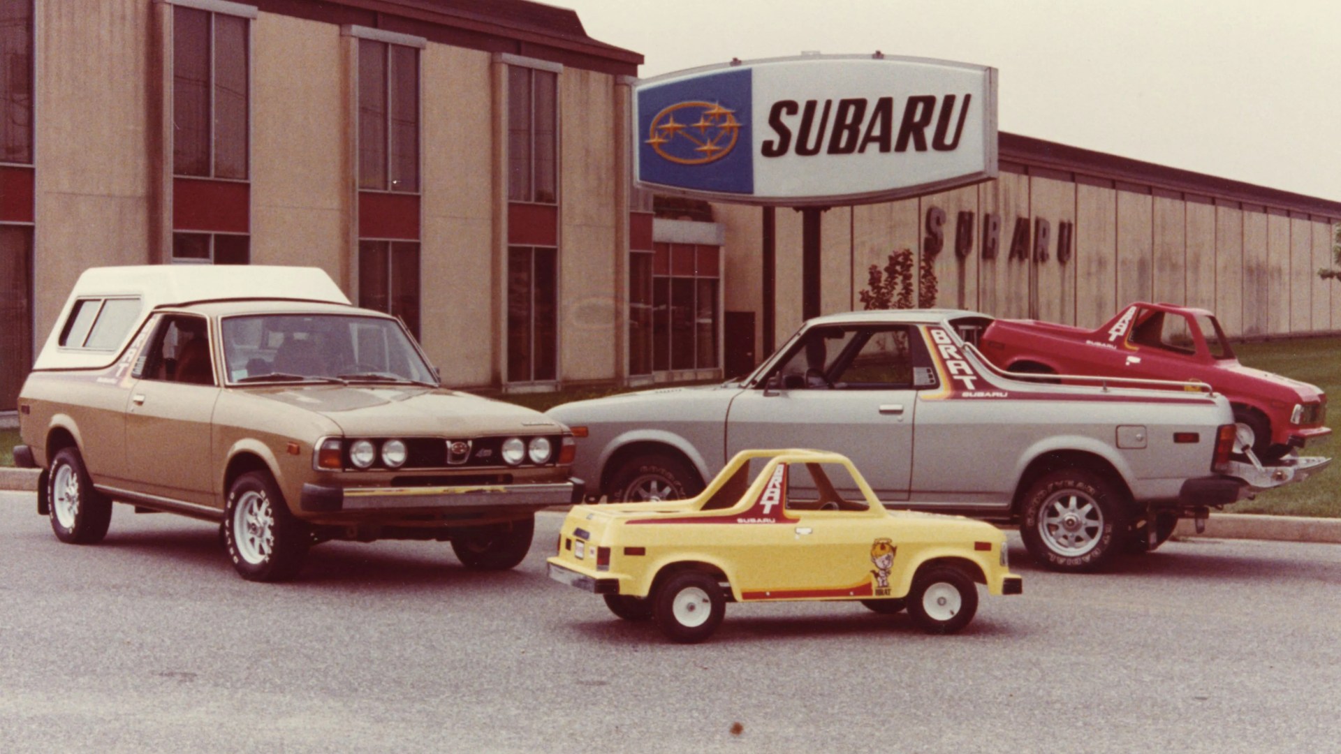 Four vintage Subaru BRAT pickup trucks in beige, silver, red, and yellow parked in front of a Subaru dealership building with a large Subaru sign.