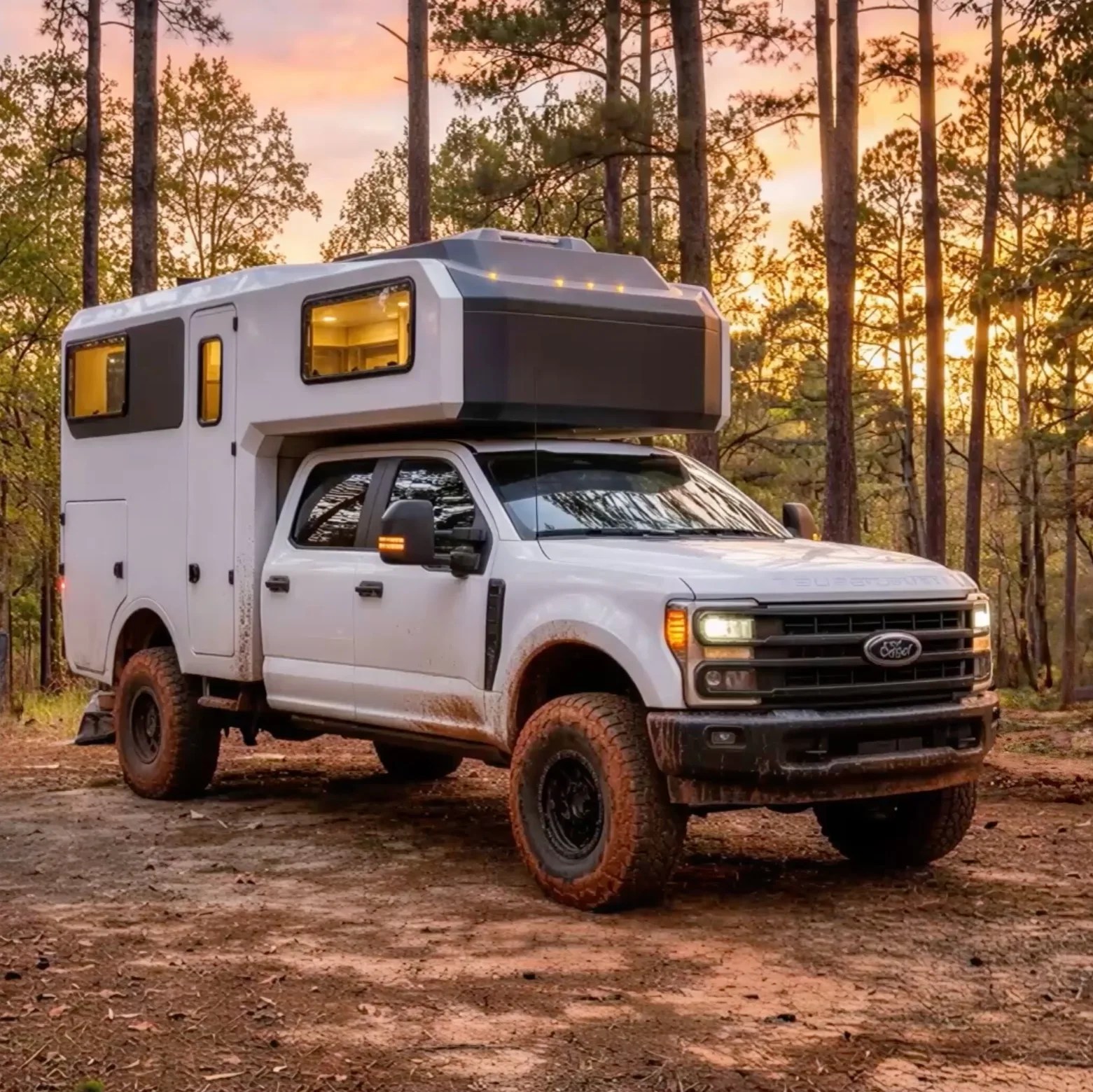 White Ford pickup truck with a camper attachment parked on a dirt path in a forest at sunset.