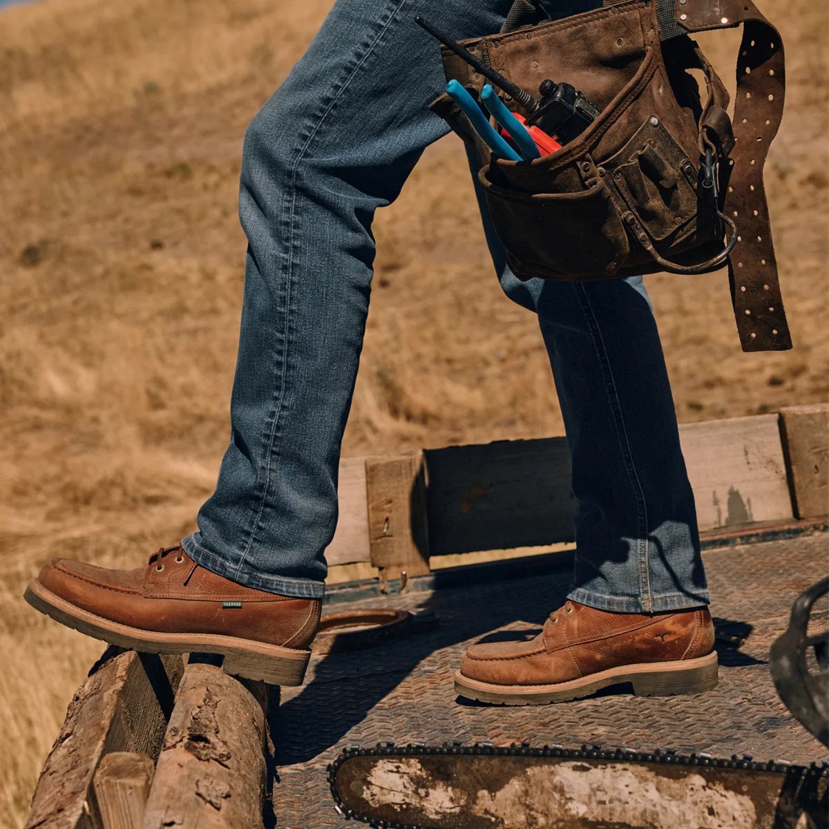Brown leather work boots worn with blue jeans standing on wooden logs outdoors.