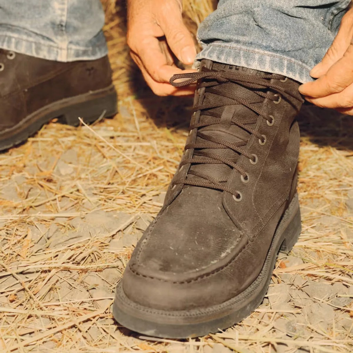 Brown suede lace-up boot worn with blue jeans on dry grass and dirt ground.