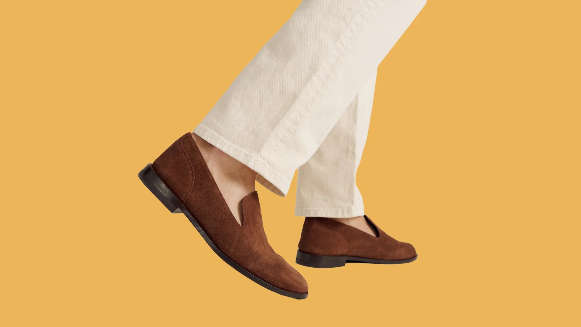 Brown suede slip-on loafers worn with off-white pants against a mustard yellow background.