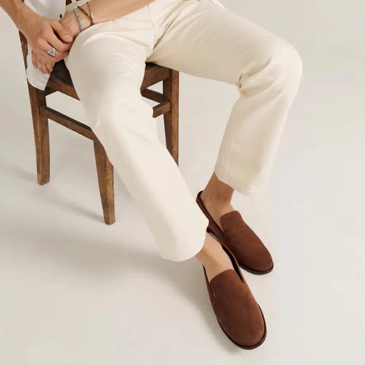Brown suede slip-on loafers worn with off-white pants while sitting on a wooden chair.