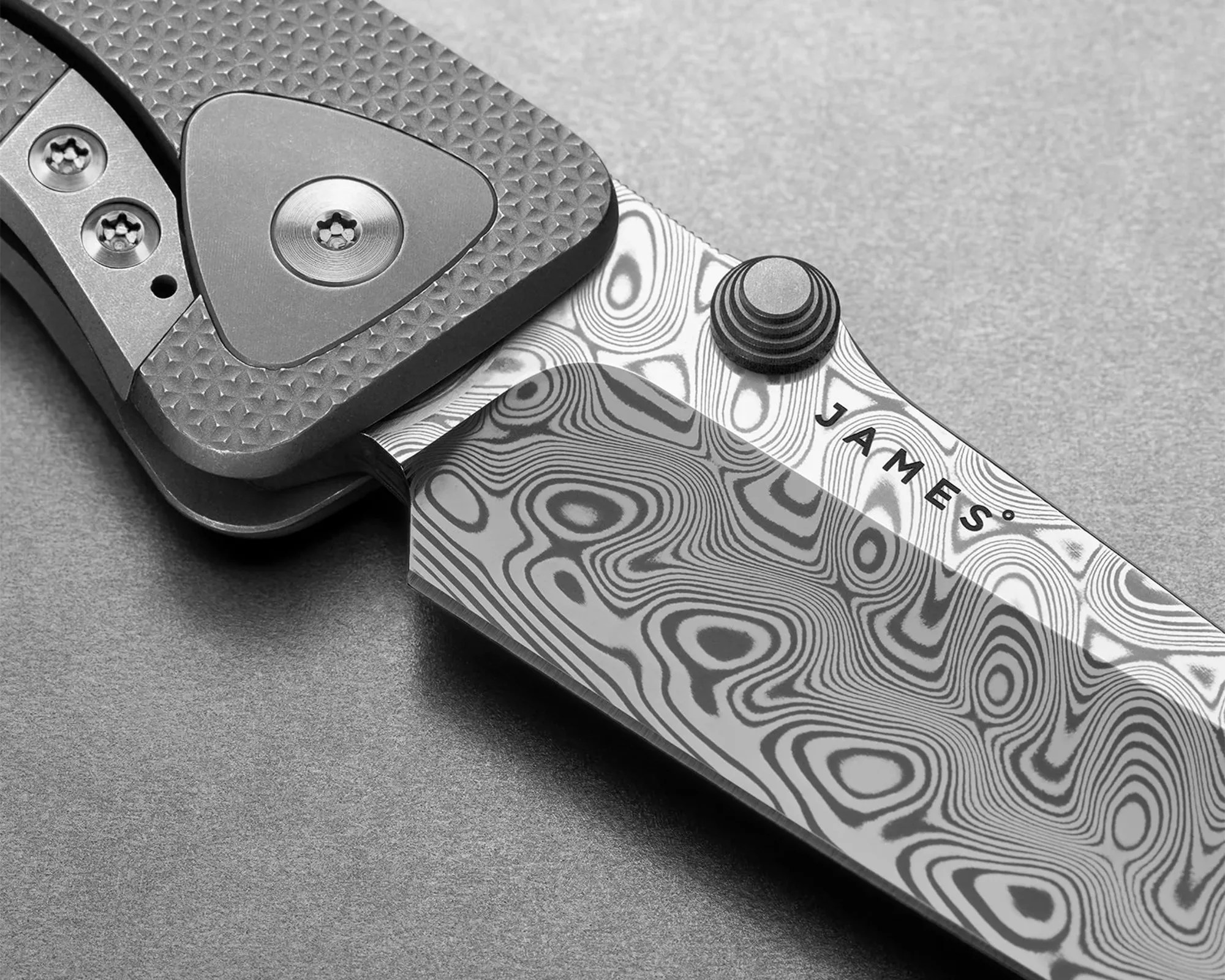 Close-up of a folding knife with a patterned Damascus steel blade engraved with "JAMES" and a textured handle with screws.