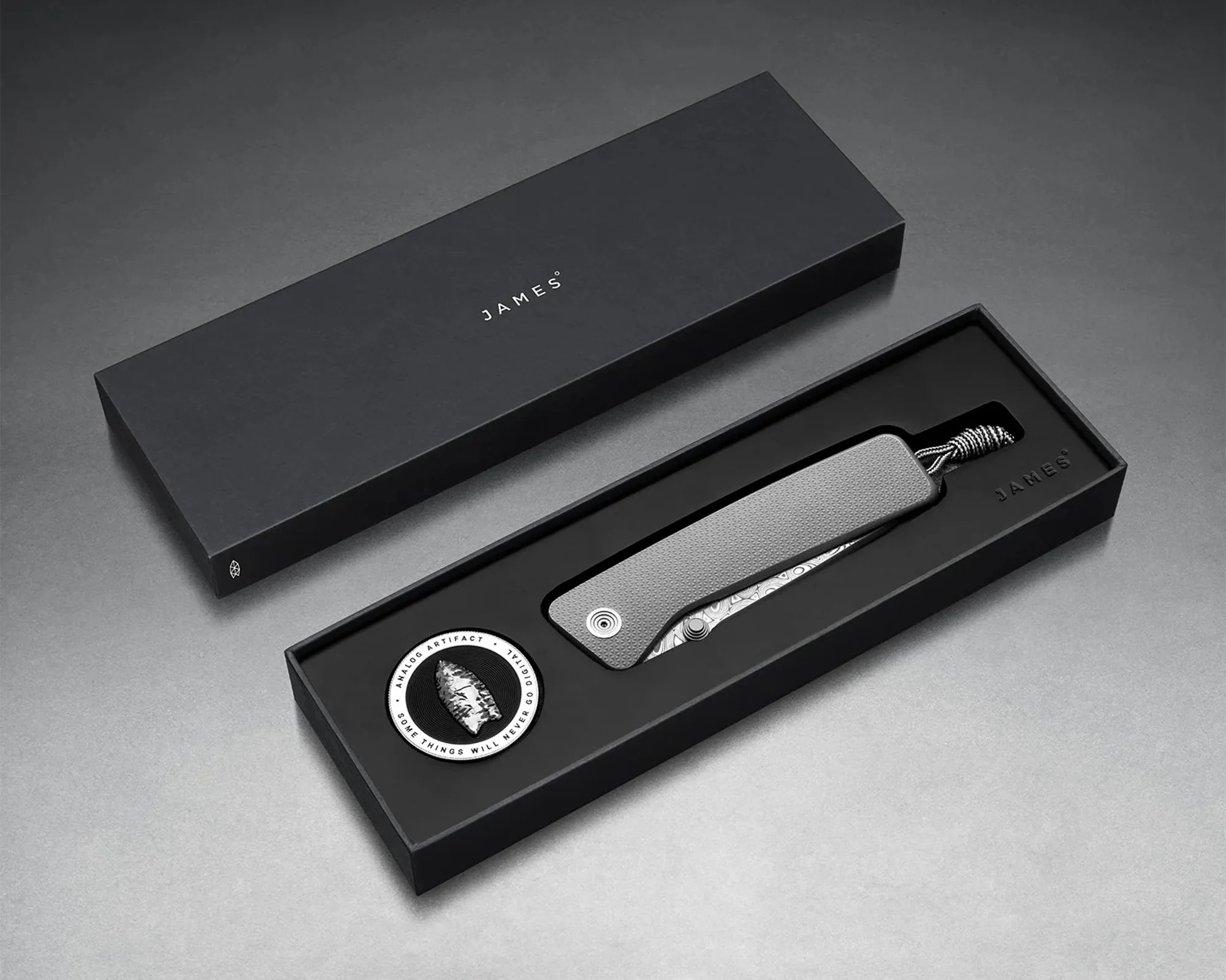 Black textured folding knife with patterned blade and braided lanyard in a black box with a round patch.
