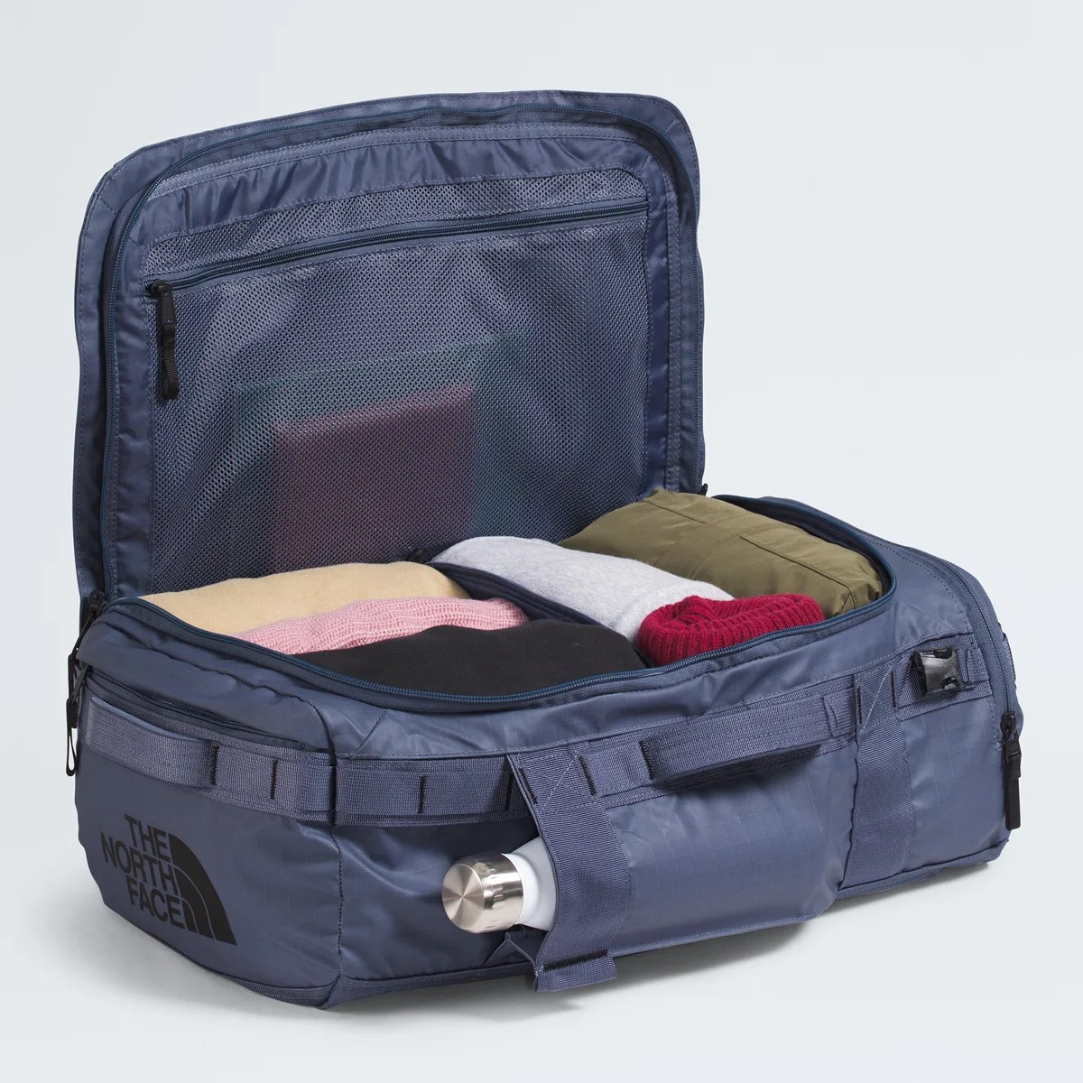 Blue rectangular soft-sided suitcase with The North Face logo, packed with folded clothes and a water bottle in a side pocket.