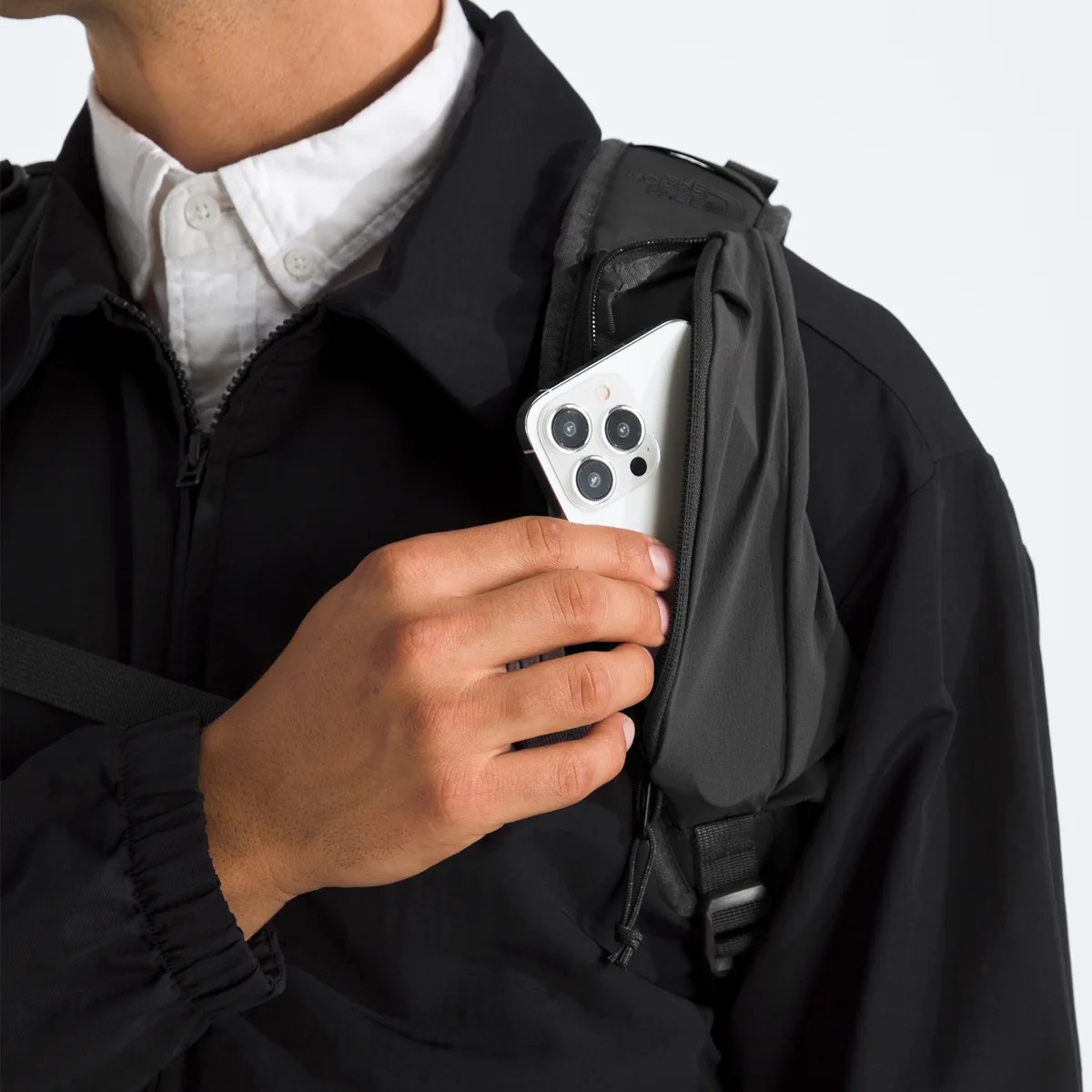 Hand placing a white smartphone with triple camera into a black zippered backpack pocket worn over a black jacket.