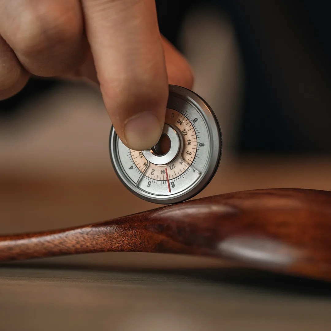 Hand holding a small circular metal gauge measuring the curvature of a wooden spoon.