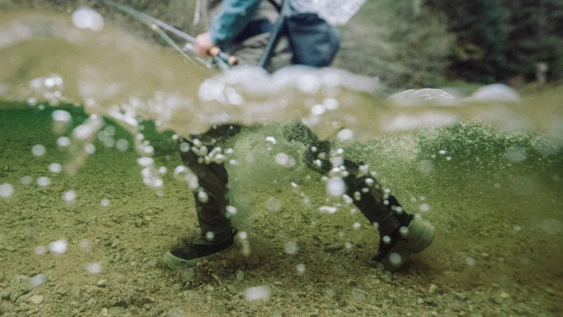 Person wearing dark waterproof boots standing underwater on a rocky riverbed with bubbles rising.