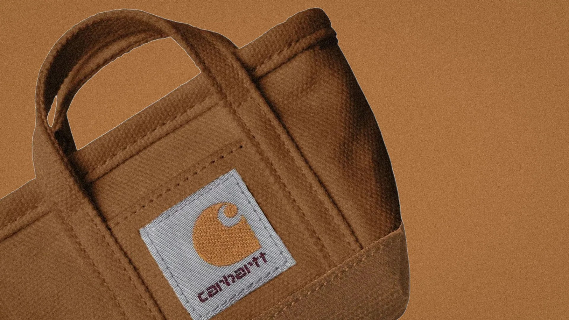 Brown Carhartt canvas tote bag with white and orange logo patch on a brown background.