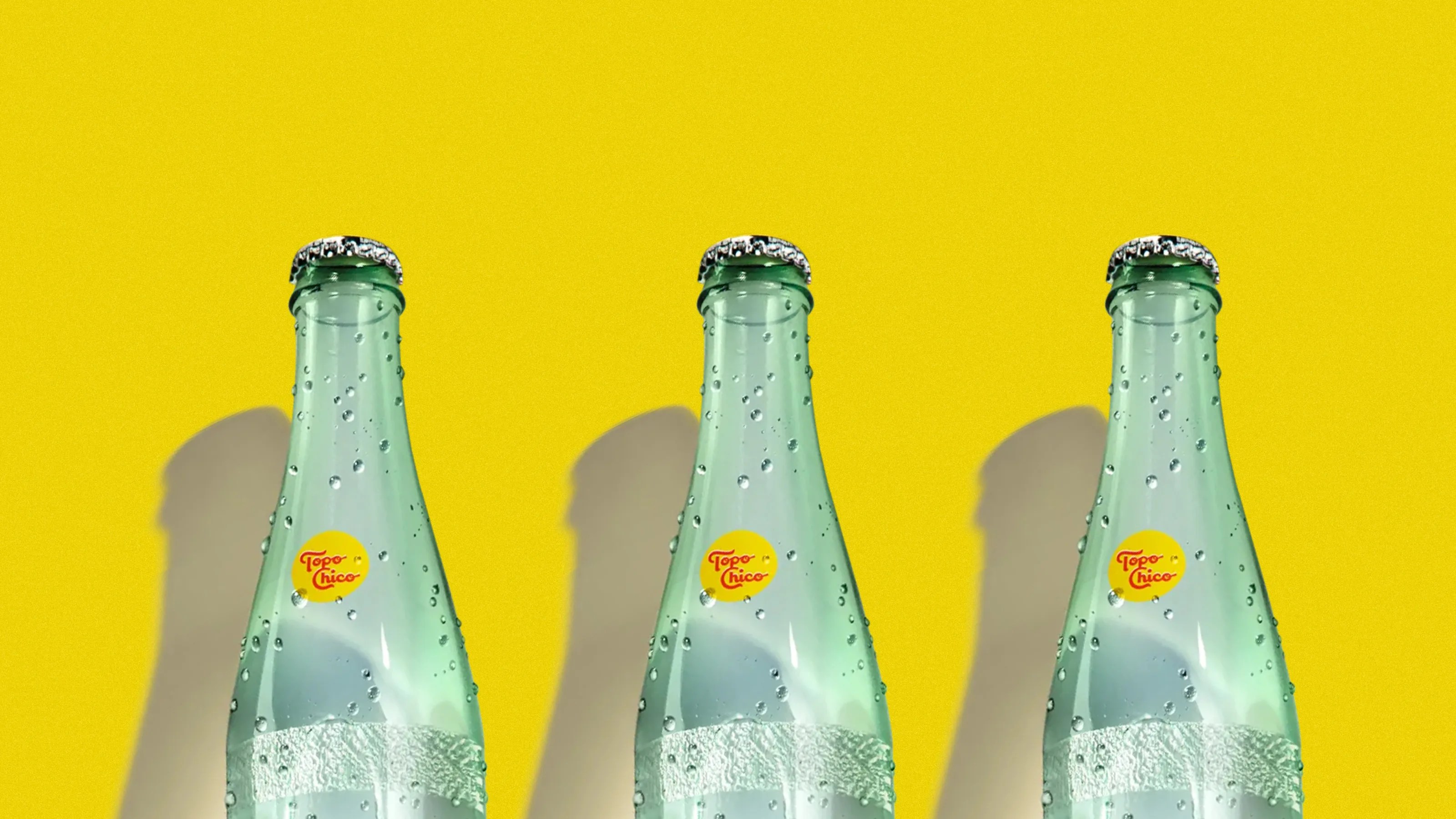 Three green glass Topo Chico bottles with condensation droplets against a bright yellow background.
