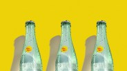 Three green glass Topo Chico bottles with condensation droplets against a bright yellow background.