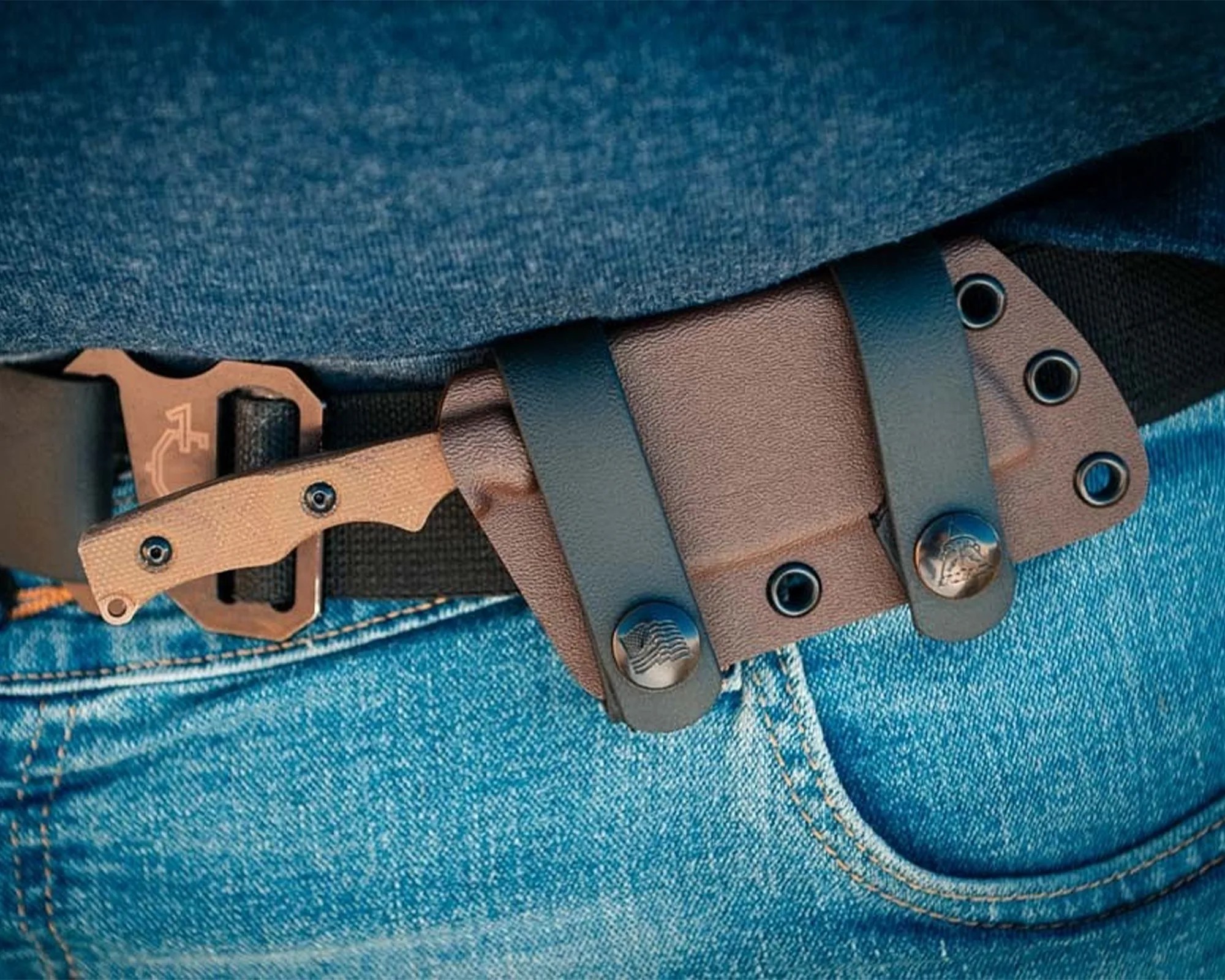 Tan-handled fixed blade knife in a brown leather belt sheath worn on a black belt with a metal buckle over blue jeans.