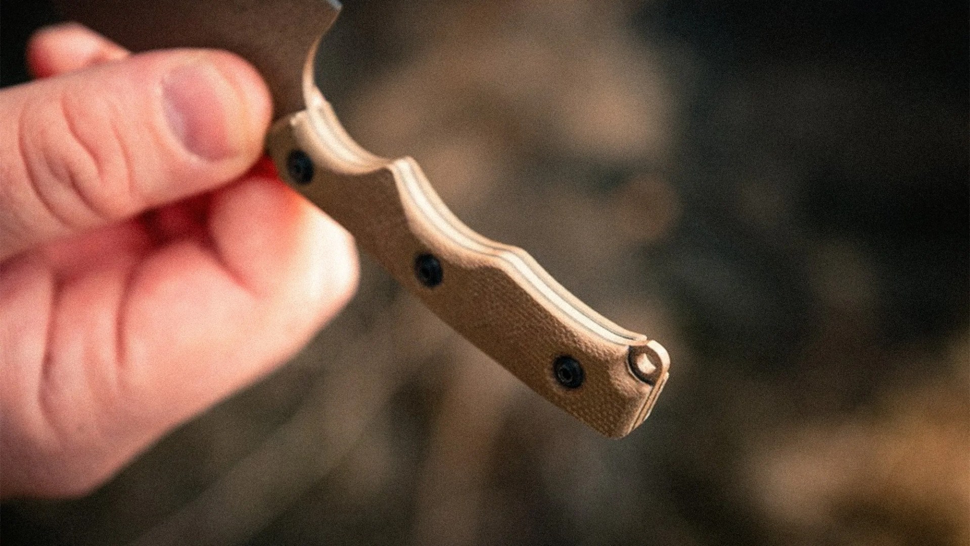 Hand holding a tan textured knife handle with black screws and a lanyard hole.