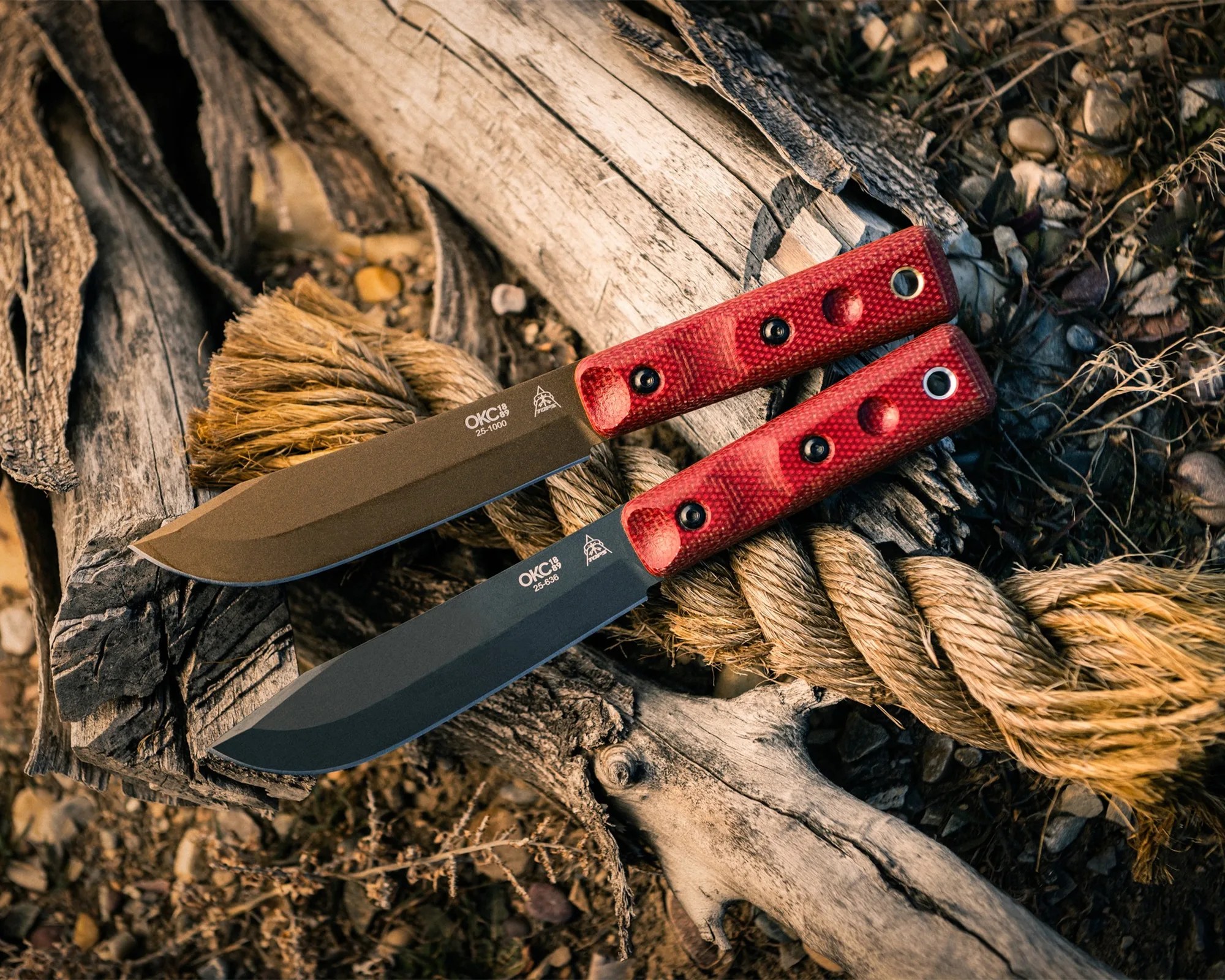 Two fixed-blade knives with red textured handles resting on a thick rope and weathered wood.