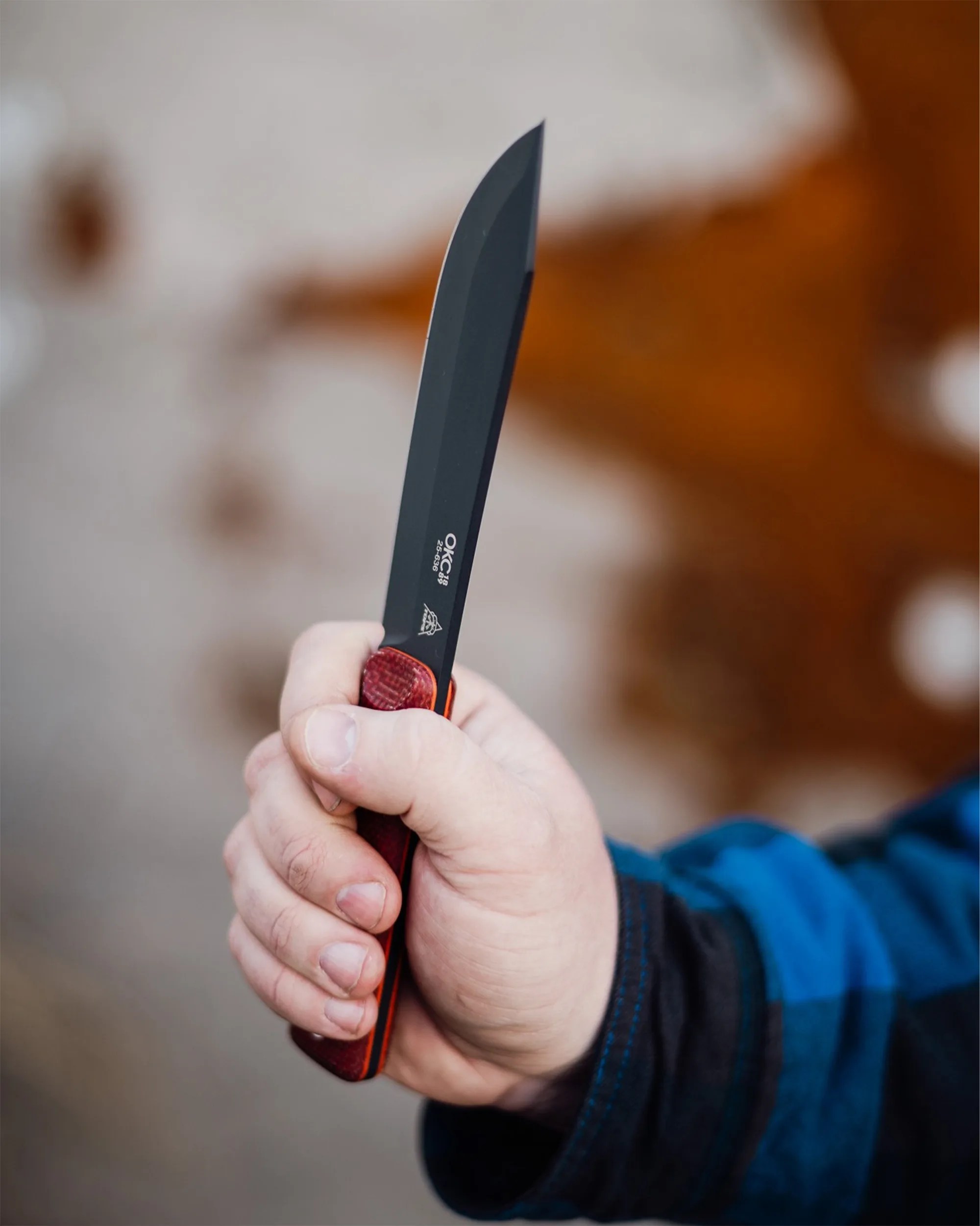 Hand holding a black blade knife with a red textured handle and visible brand markings on the blade.
