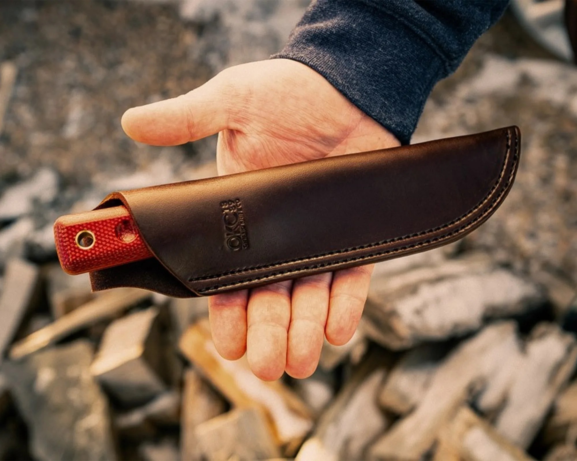 Hand holding a brown leather knife sheath with a red handle partially visible inside.
