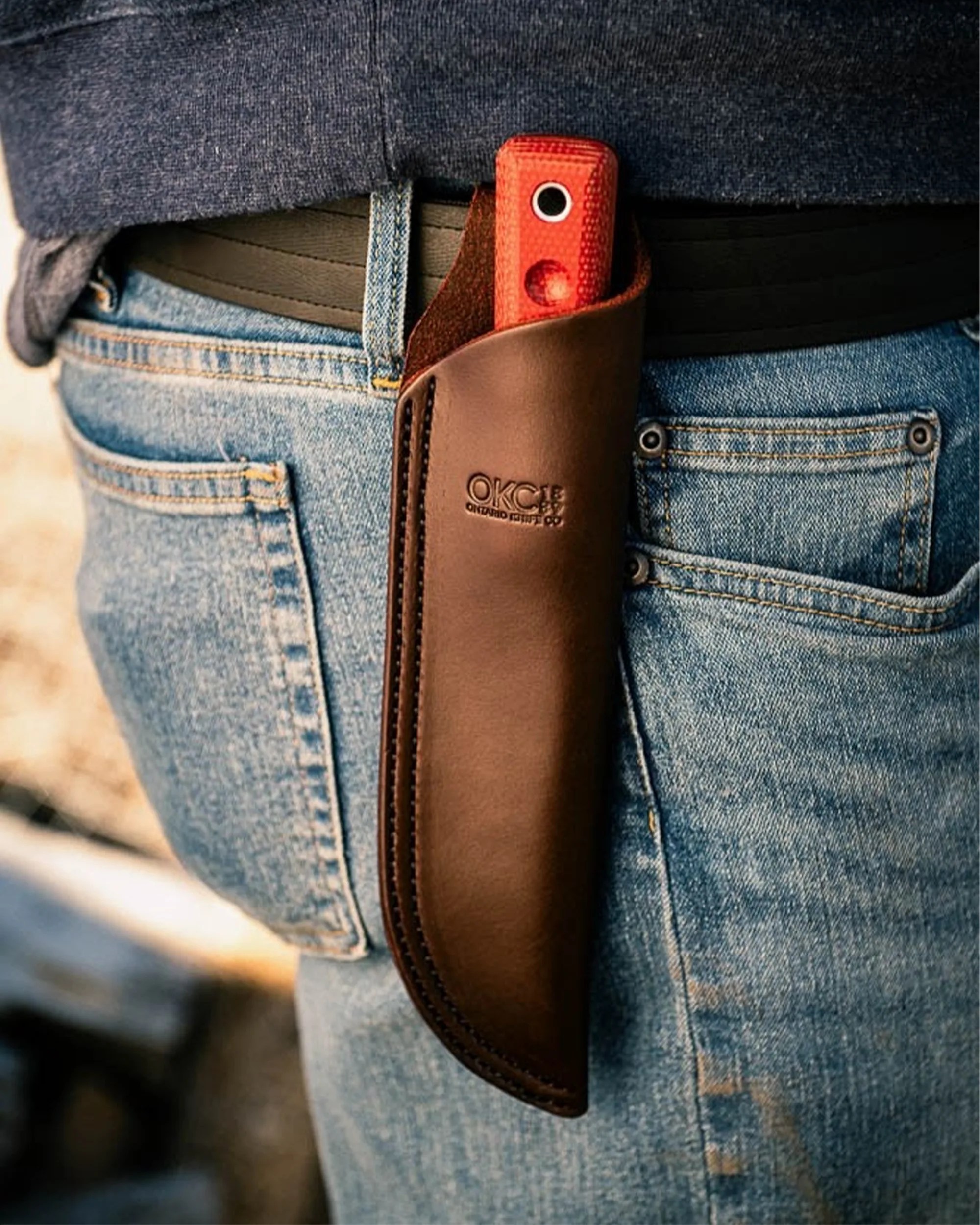 Brown leather knife sheath with embossed OKC logo holding a knife with a red textured handle worn on a black belt over blue jeans.