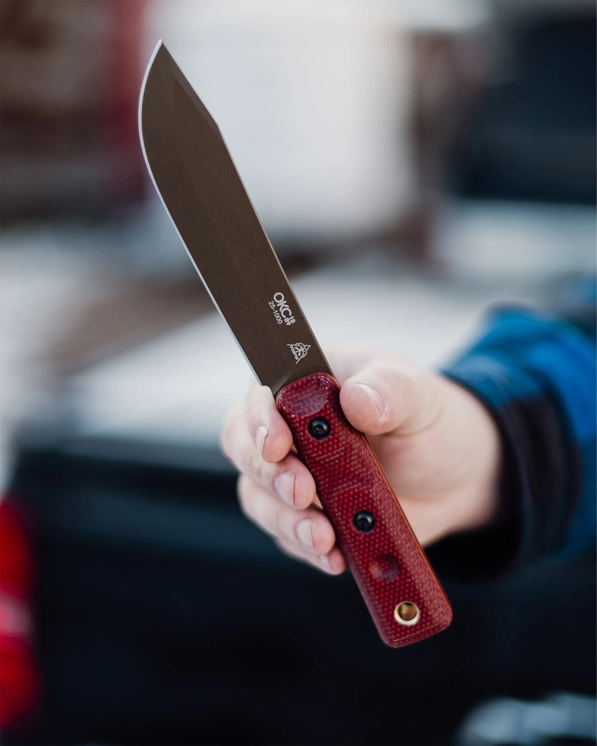 Hand holding a fixed-blade knife with a dark blade and red textured handle.