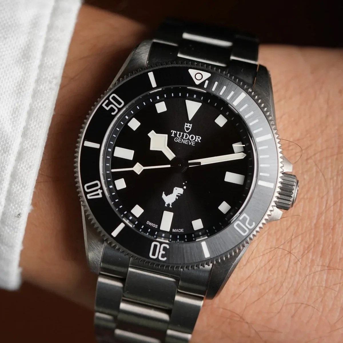Black Tudor wristwatch with white markers and a pixelated dinosaur icon on a metal bracelet worn on a wrist.