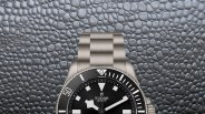 Close-up of a Tudor wristwatch with a black dial, white markers, and a stainless steel bracelet on a textured gray surface.
