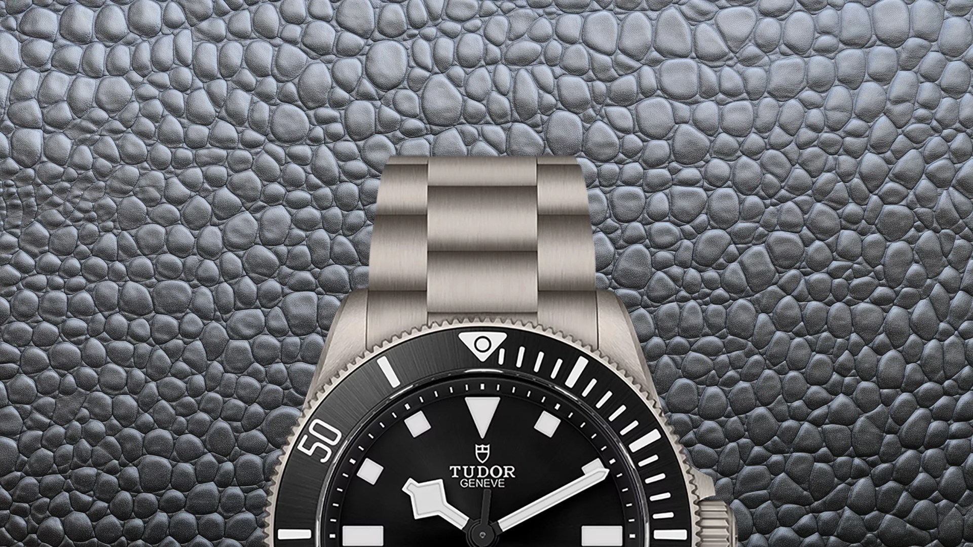Close-up of a Tudor wristwatch with a black dial, white markers, and a stainless steel bracelet on a textured gray surface.
