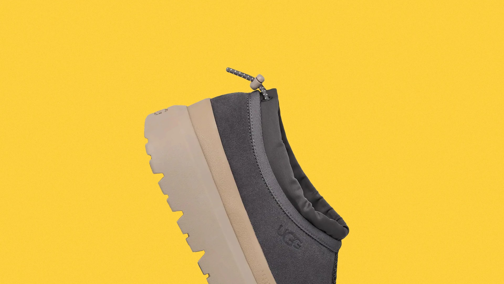 Dark gray UGG slip-on shoe with beige chunky sole and adjustable cord on a yellow background.
