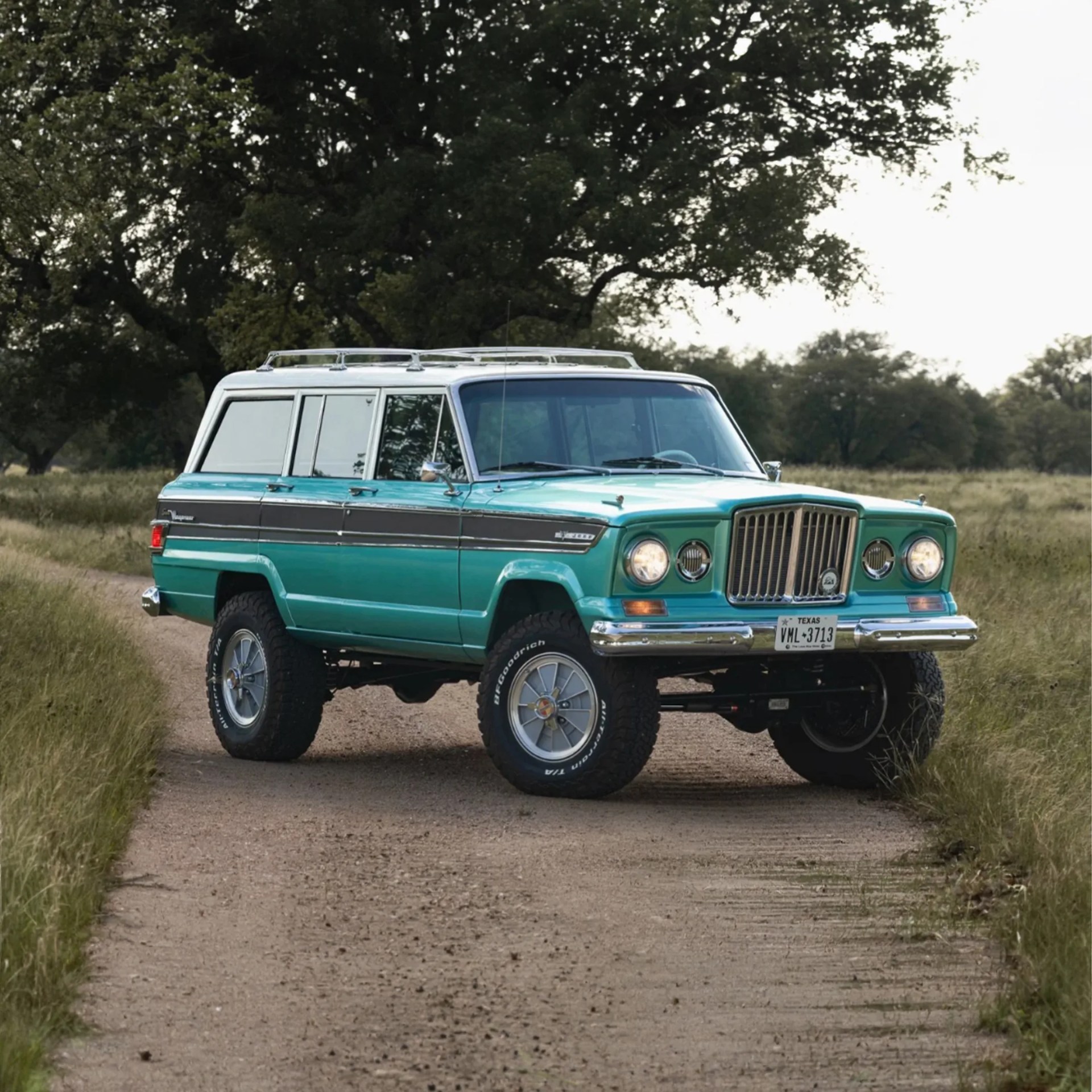 Teal and white vintage Jeep Wagoneer with off-road tires parked on a dirt road surrounded by grass and trees.