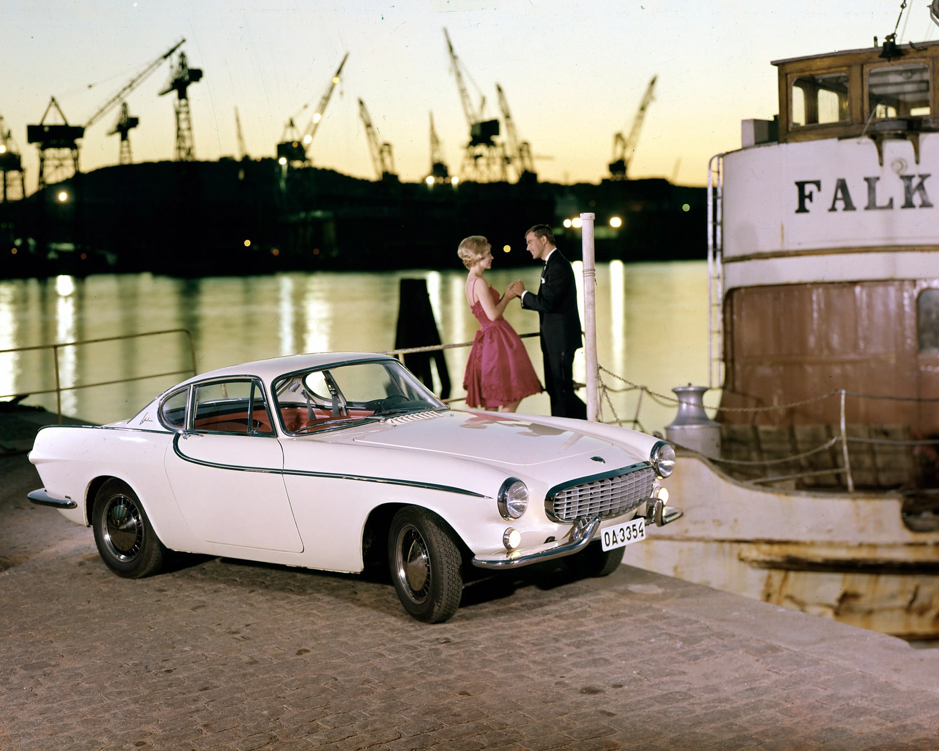 White classic Volvo P1800 coupe parked on a dock with a couple dancing near a boat named "FALK" at sunset.